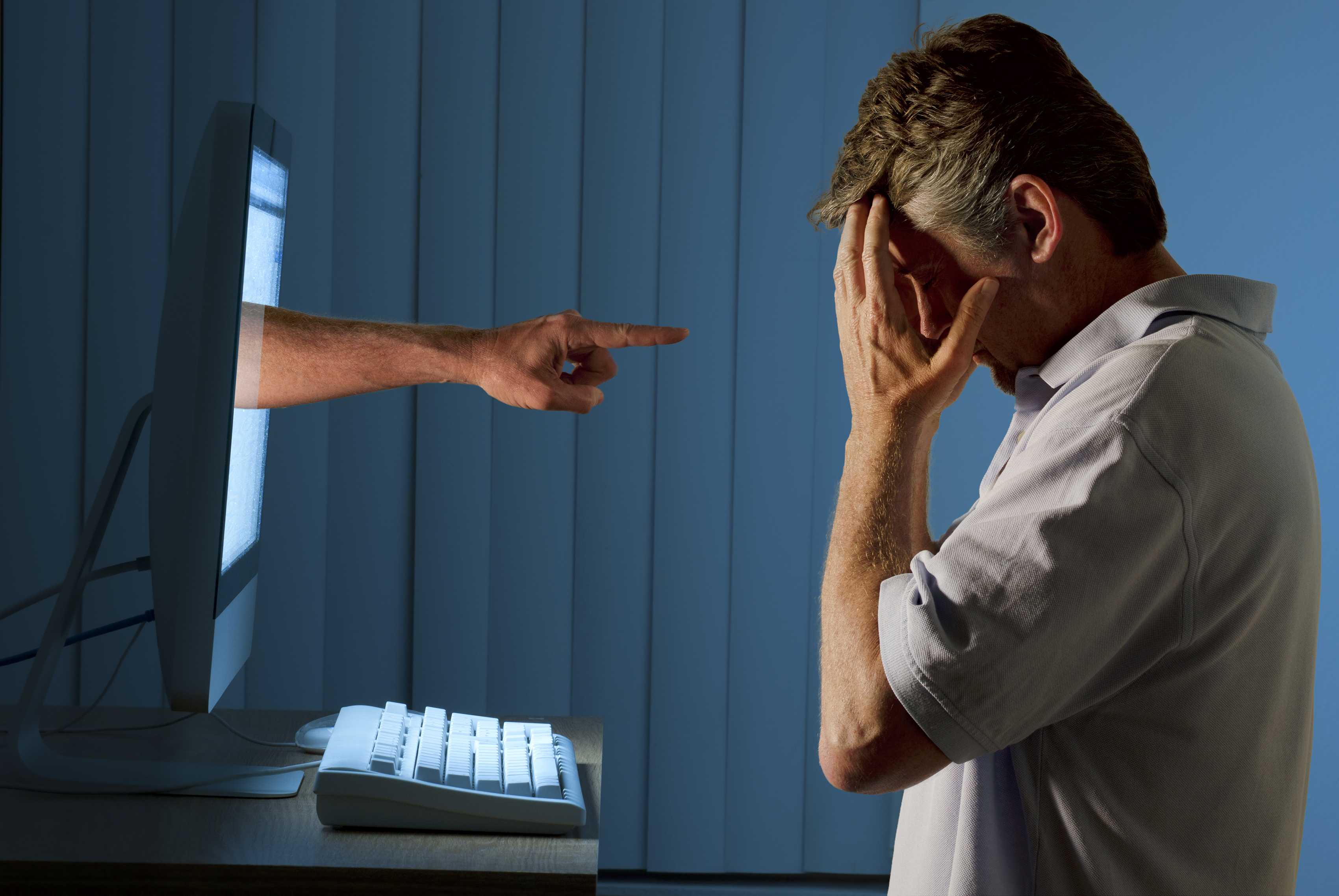 Severely distraught young man sitting in front of a computer with a judgmental hand pointing at him from within the computer monitor