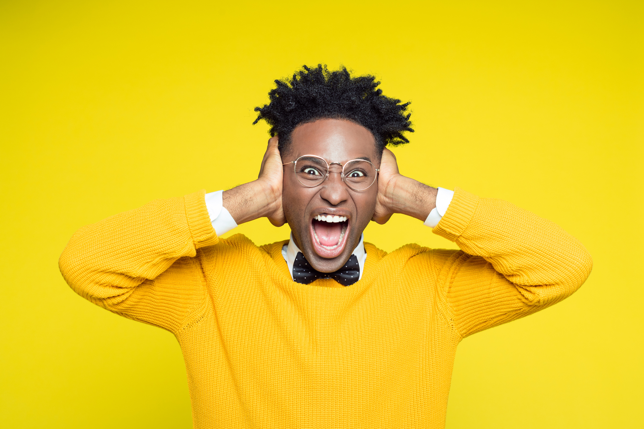 Portrait of angry nerdy young afro american man wearing yellow sweater and black bow tie shouting at the camera against yellow background.