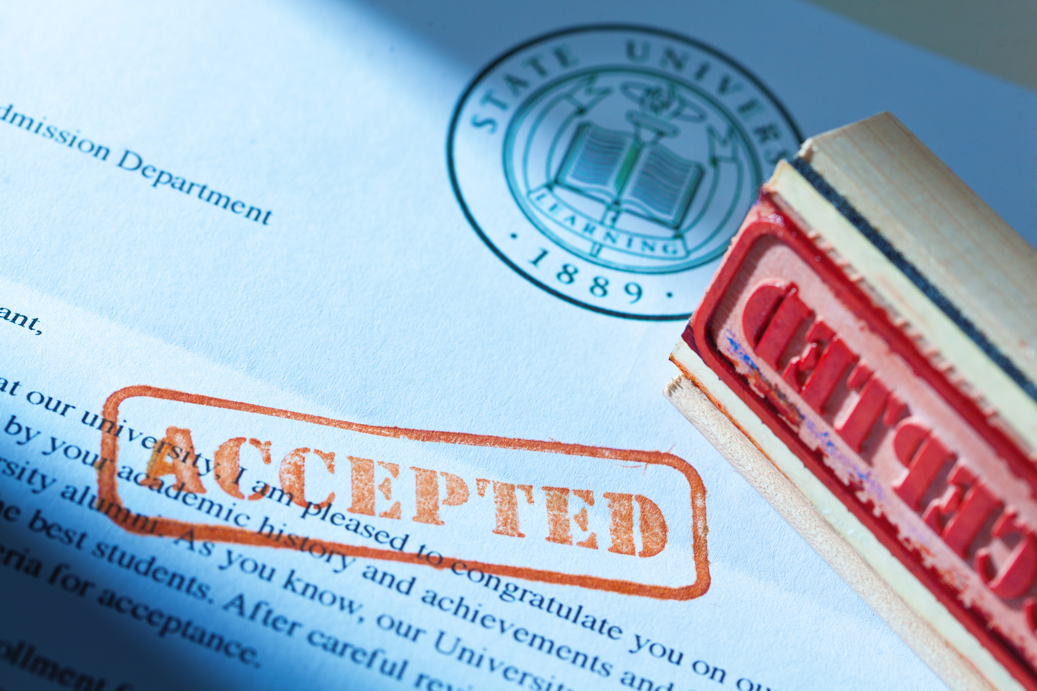 An acceptance letter from a university application. An university application form and the letter of acceptance with a red rubber stamp of "Accepted" on a table top still life. Photographed close-up in horizontal format with selected focus on the rubber stamp impression.