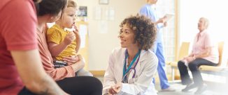 A young family sits talking to the doctor. The toddler is sat on his mother’s knee as the doctor kneels down to talk to him