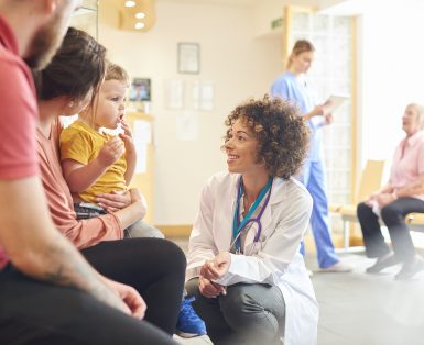 A young family sits talking to the doctor. The toddler is sat on his mother’s knee as the doctor kneels down to talk to him