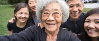 A fit ,senior, Asian Grandmother smiles with her children and mixed-ethnic grandchildren while posing for a selfie in a park garden. Real, three generation family including a senior woman, mature adult son and daughter and teenage grand-daughters. An assisted living facility can be seen in the background. Camera point of view.