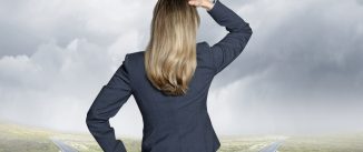 A businesswoman places her head on hand on her head as she stands at a fork in the road while looking ahead toward the horizon as the two roads disappear into the haze and mist.