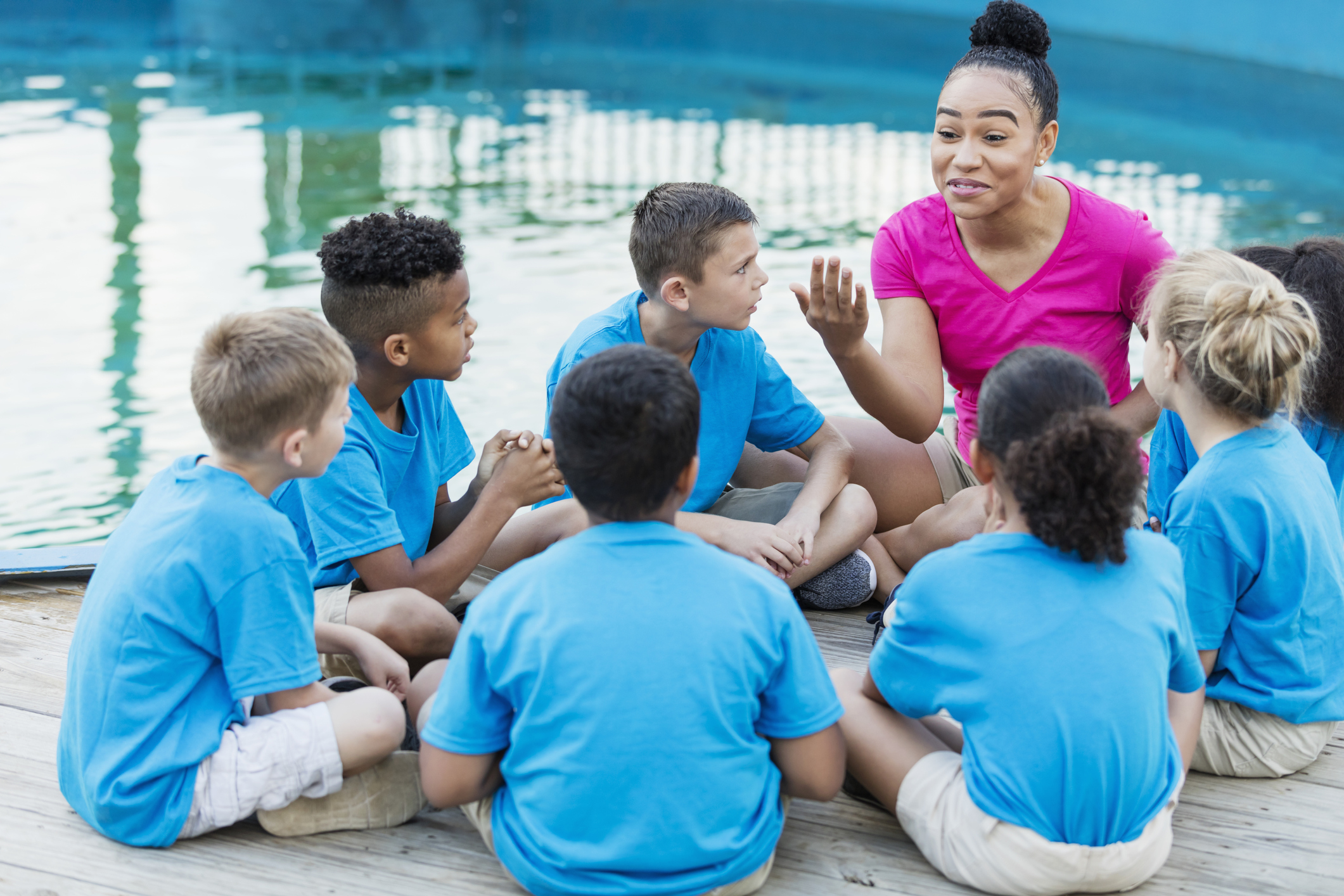 A group of multi-ethnic children on a field trip to a marine park. They are sitting in a circle by a pool, listening intently to a young African-American woman telling a story.