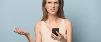 Frustrated young woman in glasses isolated on blue studio background hold smartphone