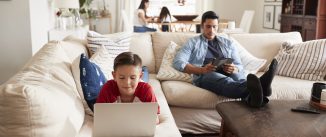 Pre-teen boy lying on sofa using laptop, dad sitting with tablet, mum and sister in the background