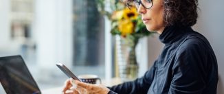 Mature businesswoman sitting at cafe looking at her cell phone while working on laptop computer. Woman reading text message at coffee shop.