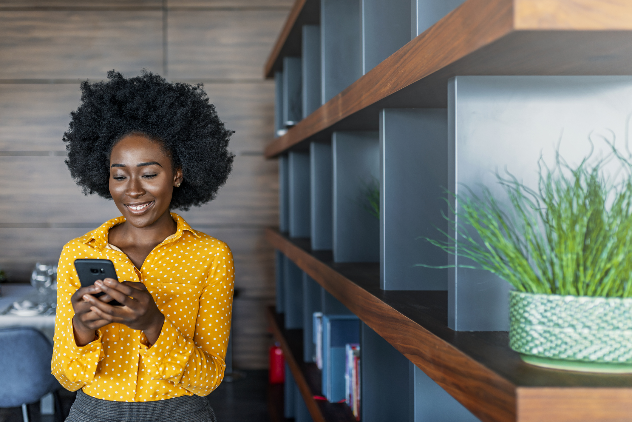 Quick message to friend. Attractive young African woman holding smart phone and looking at it while standing indoors.