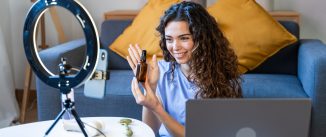 Curly-haired woman showing a skincare oil bottle while recording a video with a laptop and ring light